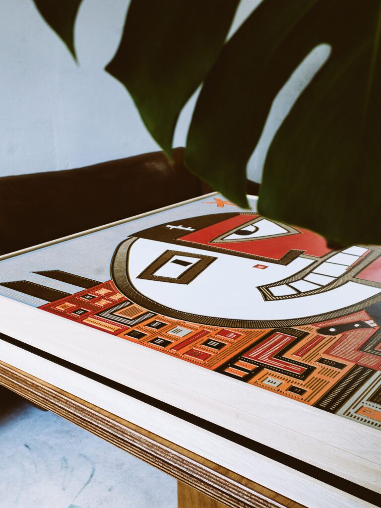 Backstabbed Boy by Zolo Kis — angled view of the framed red and white face on a wooden table beside a plant