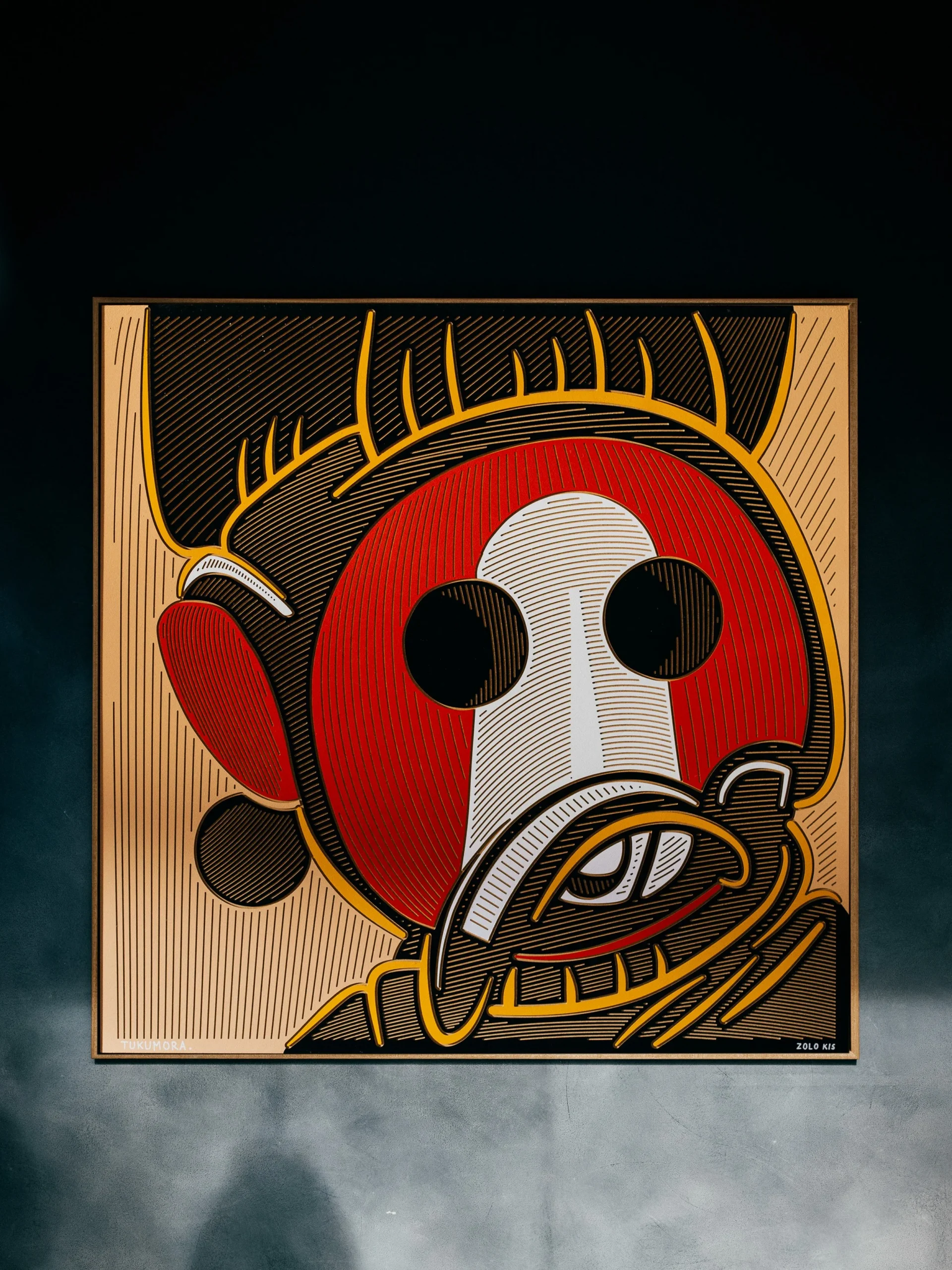 Tukumora by Zolo Kis — framed red and white warrior face with round black eyes and yellow outlines on a dark wall